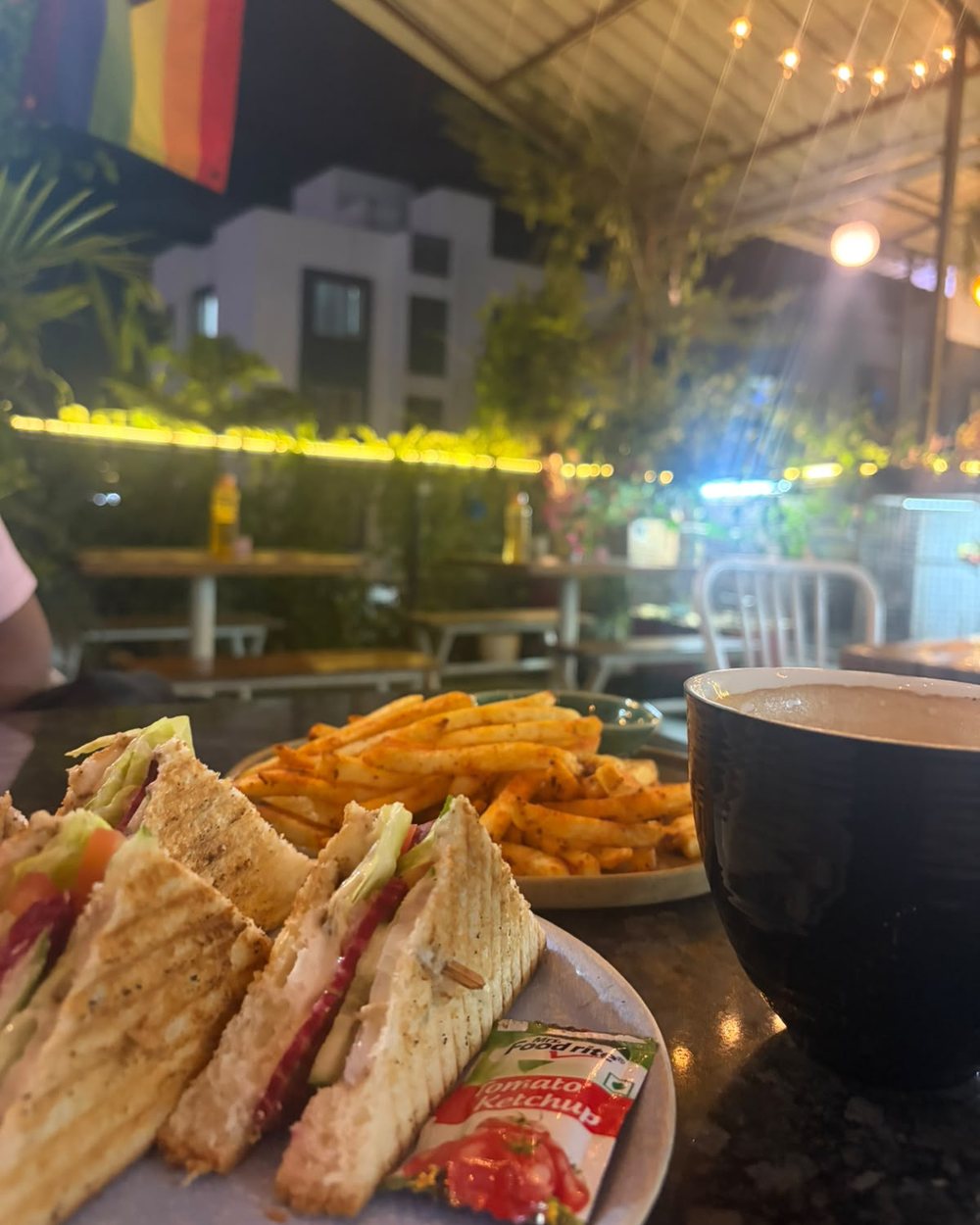 A grilled sandwich, fries and coffee on a table under fairy lights at night